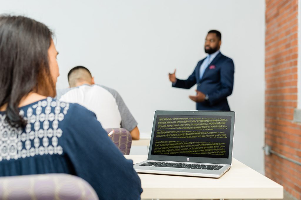 Man presents to an audience as woman looks at her laptop